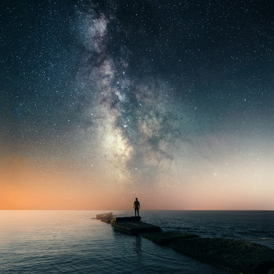 Person standing on rocks leading into the ocean, gazing at the Milky Way in a starry night sky.