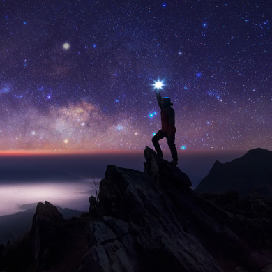 Person standing on a mountain peak at night, reaching toward a bright star under a star-filled sky.
